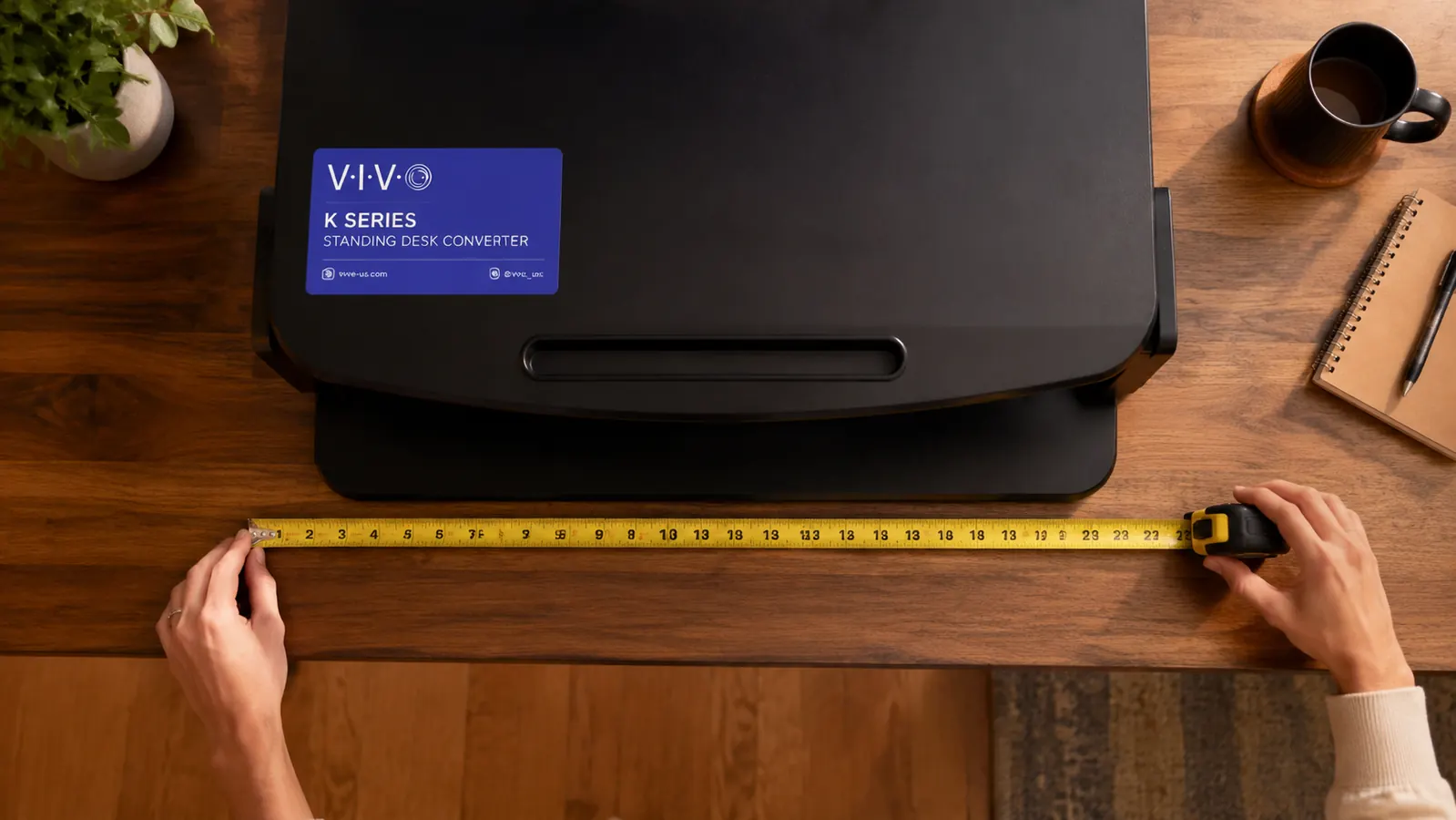Measuring tape laid across a desk surface next to a standing desk converter showing the depth it occupies